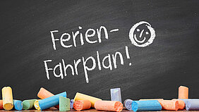Schwarze Tafel mit bunten Kreidestücken und weißer Aufschrift "Ferien-Fahrplan!"