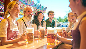 Ein Gruppe gut gelaunter Frauen und Männer in Tracht mit gefüllten Maßkrügen an einem Biertisch im Freien auf dem Volksfest.