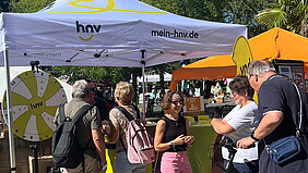HNV-Infostand mit Zelt, Infotheke und Glücksrad im Freien. Zwei interessierte Paare lassen sich vom Standpersonal beraten.