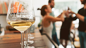 Im Vordergrund ein mit Weißwein gefülltes Erlenbacher-Weinfest-Stielglas, im Hintergrund eine Gruppe Freunde, die sich zuprosten.